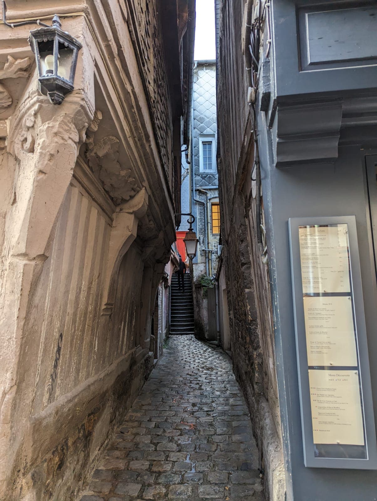 IMG-01. A narrow cobbled alley running between old buildings, the paving wet. A carved stone facade stands on the left under a wall lantern; a glazed display case holding printed sheets is mounted on the dark frontage to the right. The alley ends in a flight of steps, a figure climbing away from the camera.